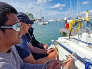 Sunny day, Ollie & Anil sitting in the cockpit practicing tying a half hitch using the rail that runs along the cockpit table. Behind is blue sea with moored boats.