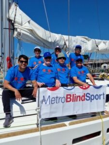 All seven crew in the Metro Blind Sport t-shirts looking out of the side of the boat, in front is a Metro Blind Sport dodger.