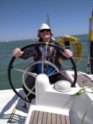 Anil, with both hands on the wheel, helming the yacht. The horzon behnd is at a slight slant showing that the boat is heeling towards port.