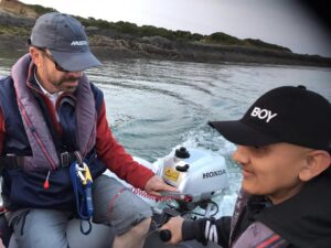 Vjay and David in the dinghy, Vjay is steering the outboard motor, David is opposite, behind is a smooth sea and the seashore.