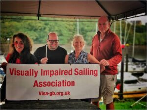 the four crew onshore taking shelter under an awning, from left to right is Malika, Andrew, Jennie and Tim, they are holding the Visa-gb banner. Behind is the green bank of trees on the far side of the marina.