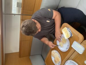 Lloyd sitting at the saloon table, knife in right hand, plate with toasted muffins and a pot of butter to his left.