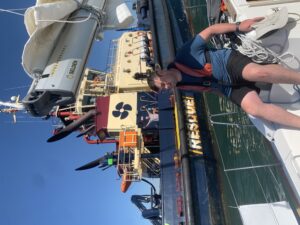 Brandon is sitting on the coach roof, behind is a very large tug with cream coloured superstructure, large funnels and a dark hull, On deck is Grandad Charlie,  wearing a grey t-shirts, he has both hands in the air .