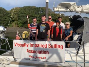 six Crew standing in the cockpit of a yacht looking out over the guard rail, there's a Visa-gb dodger in front. From left to right on the front row, Charles, Brandon, Malika, Jennie, behind Tim and Lloyd