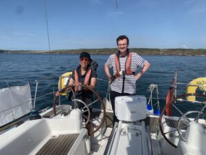 Malika on the starboardside helm with Charles standing to her left with his hands on his hips, both have life vest on, behind is a flat blue sea and above a bright blue sky.
