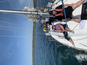 picture taken from the bow looking aft, Andrew is sitting in fron of the mast with Jennie to his right, back in the cockpit is Malika on the helm with Charles standing to her left, there is a white wake in the blue sea alongside 